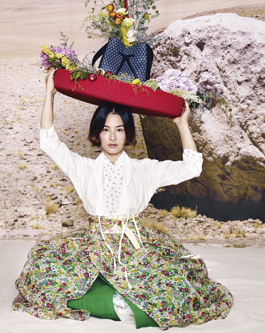 A Vogue Korea editorial look holding a flower-filled tray overhead in a garden