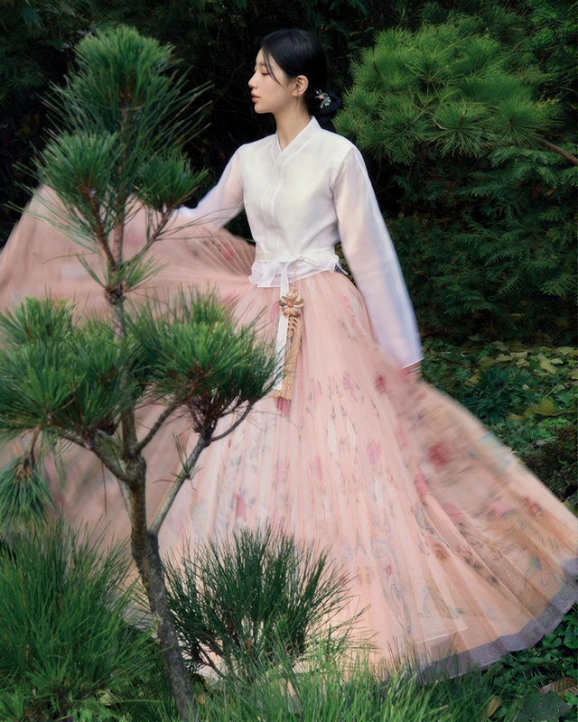 A woman wearing a pale pink hanbok among pine trees